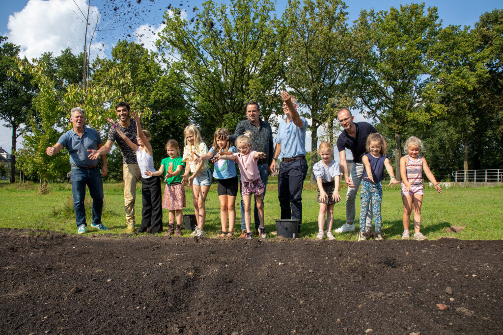 Kinderen, wethouder Steven Greving en initiatiefnemers Marko Lion en Cor Burger strooien het zaadmengel voor meer biodiversiteit uit